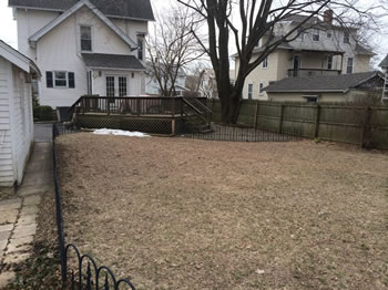 Before picture of a large backyard, void of grass, with a patio and a small fence.